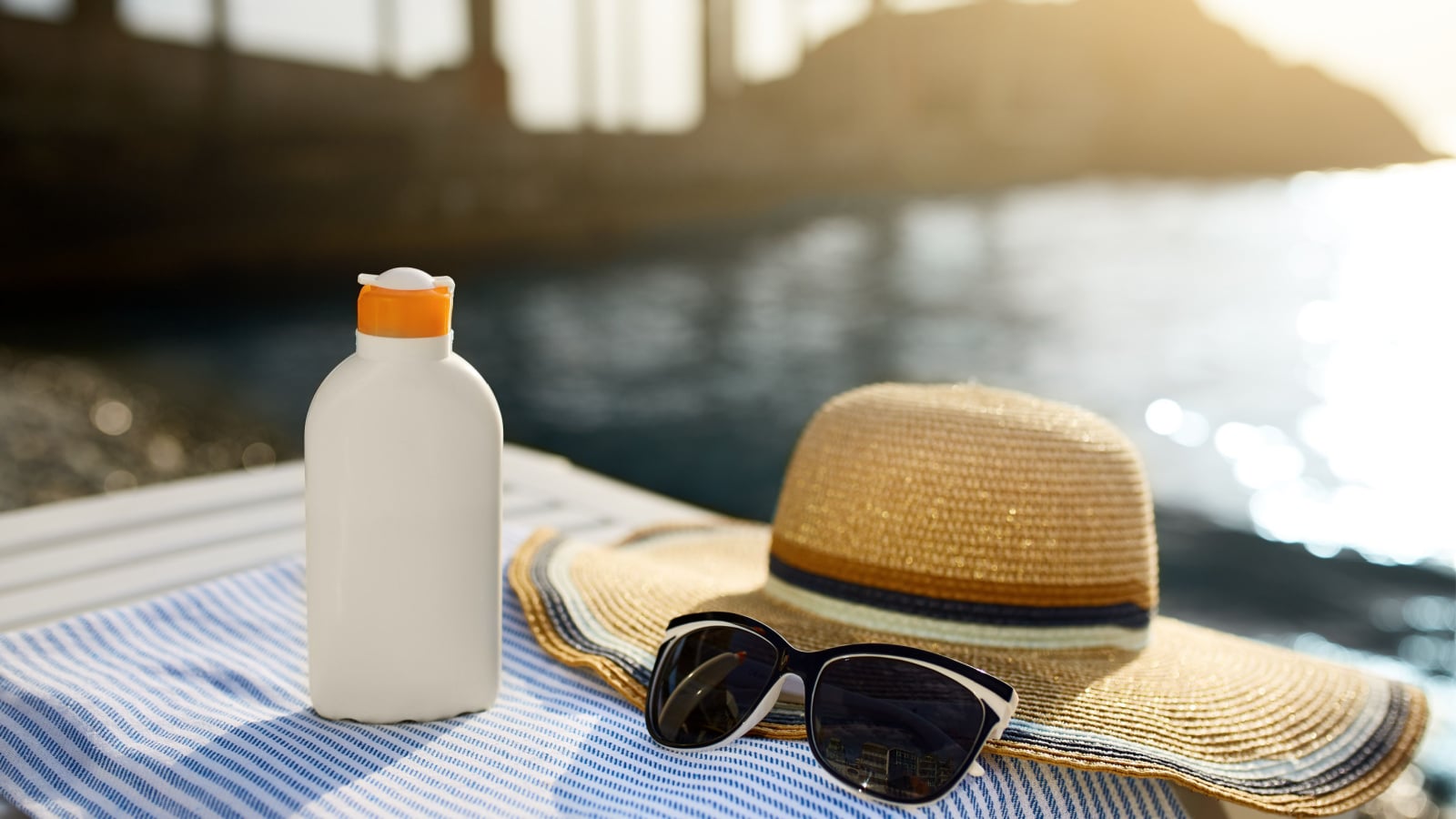 Suntan cream bottle and sunglasses on beach towel with sea shore on background. Sunscreen on deck chair outdoors on sunrise or sunset. Skin care and protection concept. Golden tan.