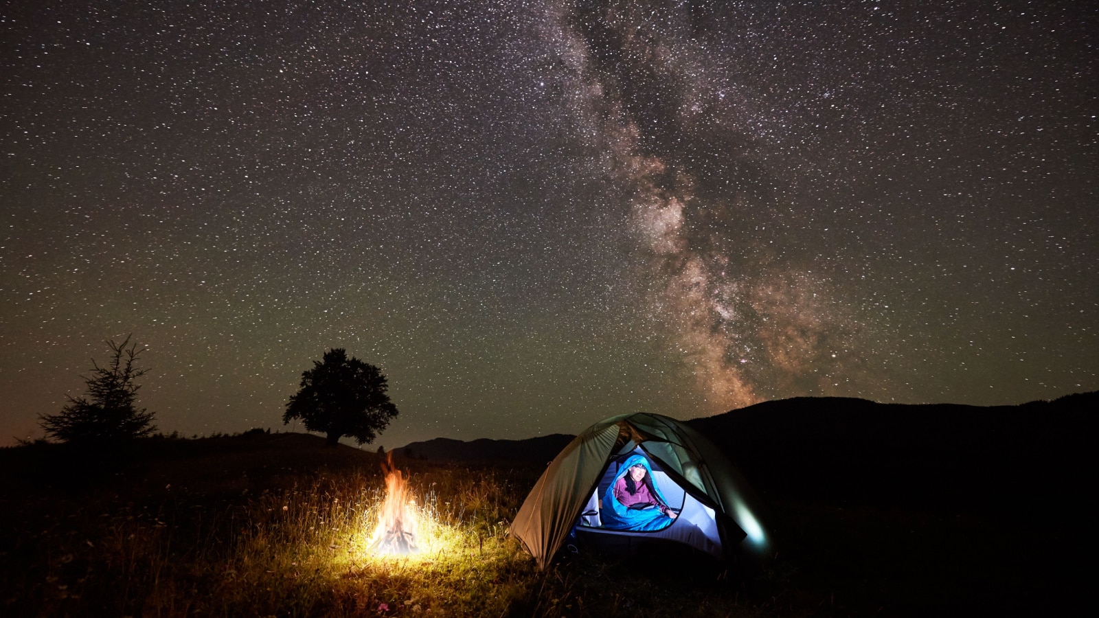 Woman hiker having a rest at night camping beside campfire in the mountains under incredible beautiful starry sky and Milky way. Girl sitting inside illuminated tent and sleeping bag. Astrophotography