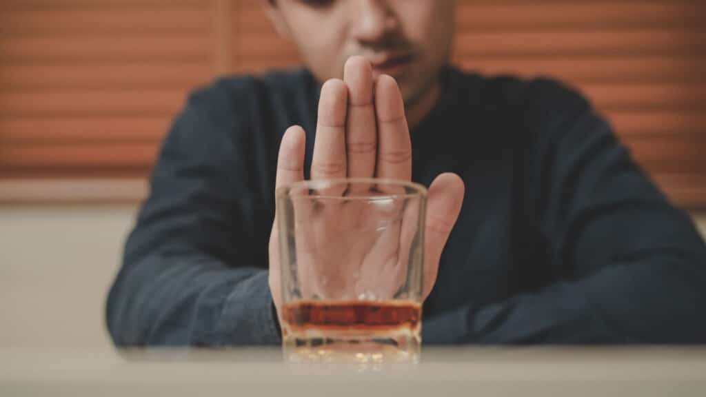 Alcoholism, sad depressed asian young man refuse, push alcoholic beverage glass, drink whiskey, sitting alone at night. Treatment of alcohol addiction, having suffer abuse problem alcoholism concept.