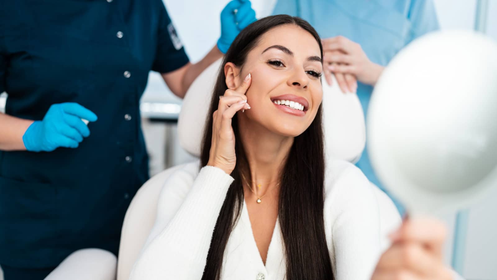 Beautiful and happy young woman sitting in medical chair and looking in the mirror. She is satisfied after successful beauty treatment with hyaluronic acid fillers or botulinum toxin injections.