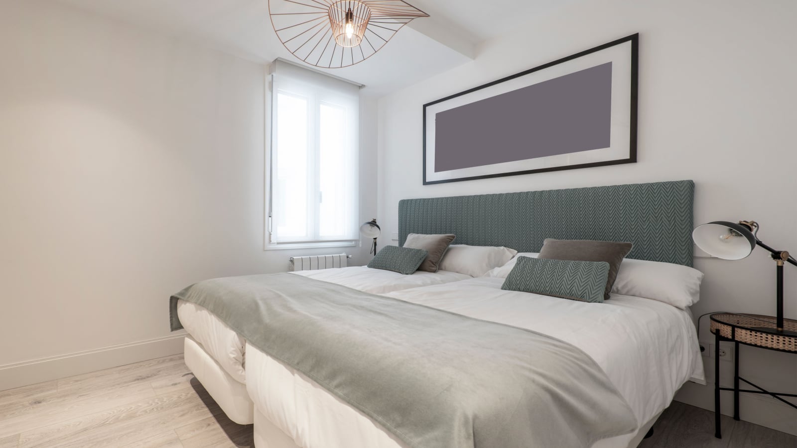 Bedroom with two twin beds pushed together, headboard upholstered in green fabric, wicker and metal bedside tables and a beautiful lamp on the ceiling