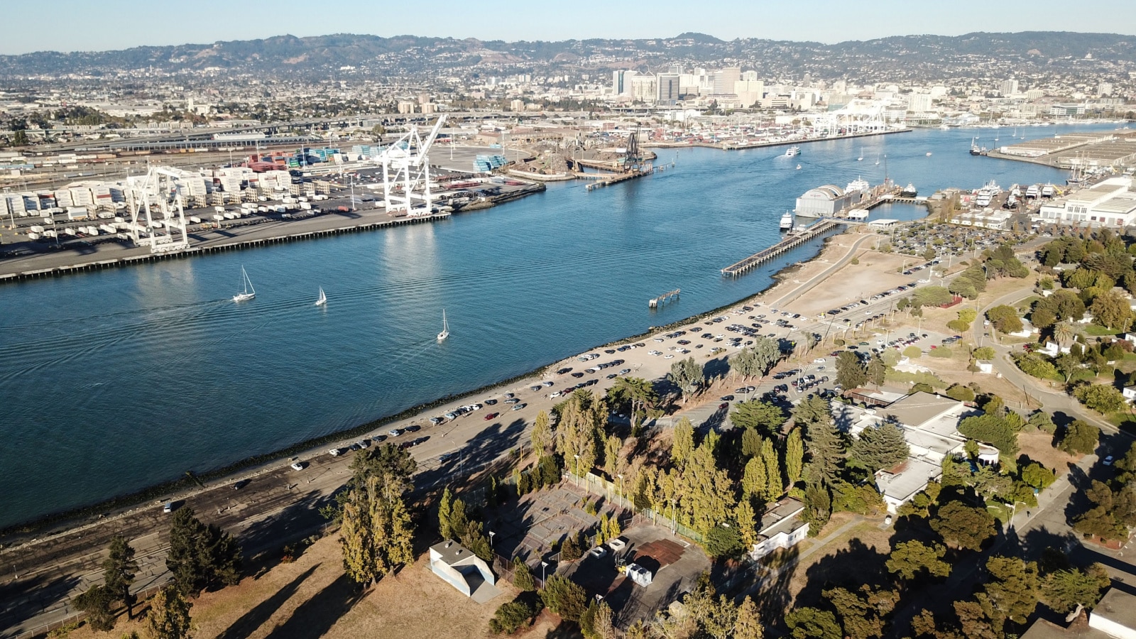 An aerial view of the busy city of Oakland, where you can see parks and residential neighborhoods on one side of a vast river, and industrial ports and the city skyline on the other side.