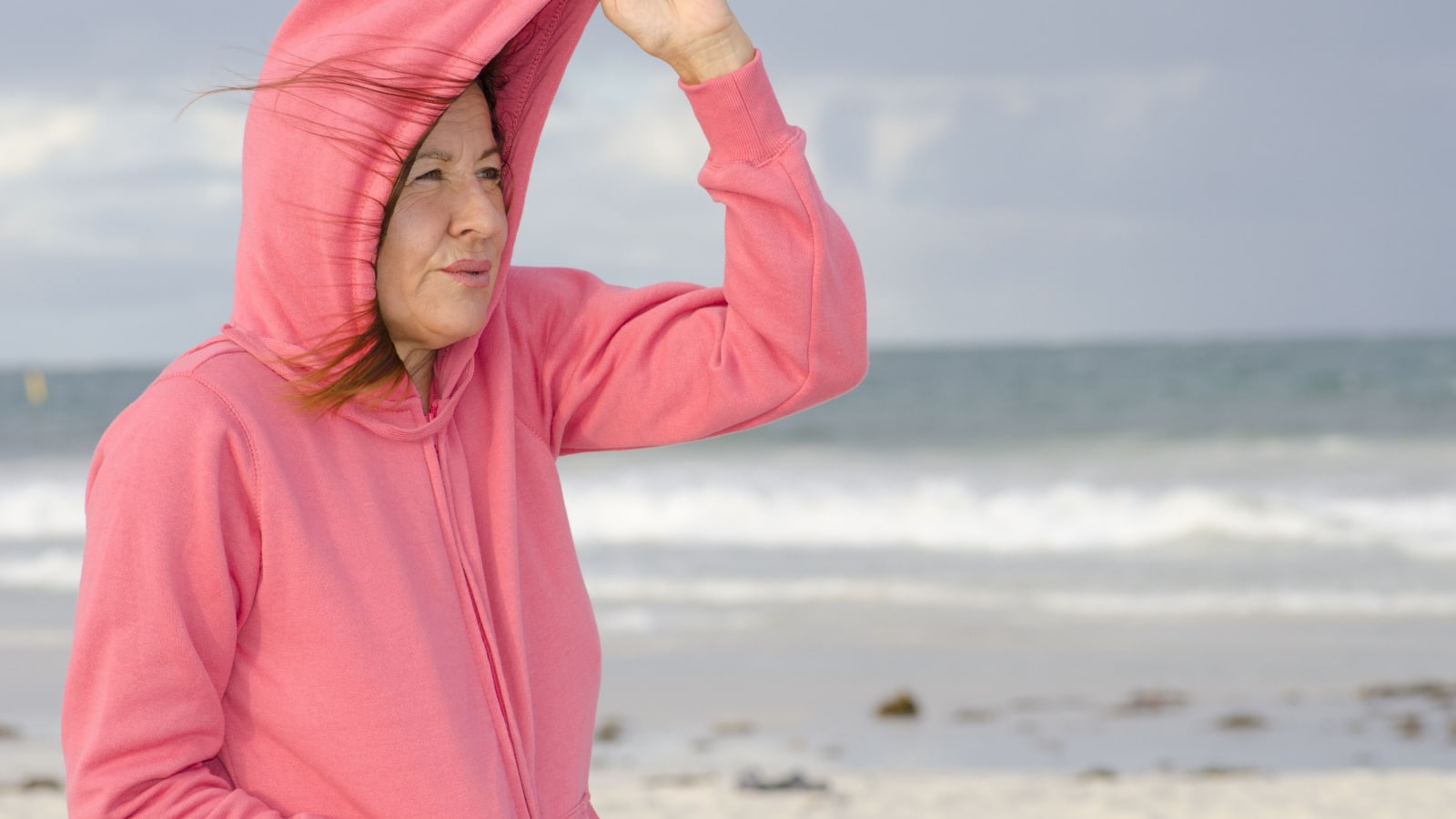 Portrait of happy attractive looking middle aged woman wearing pink sweater, standing at beach, isolated with storm clouds and ocean as background and copy space.