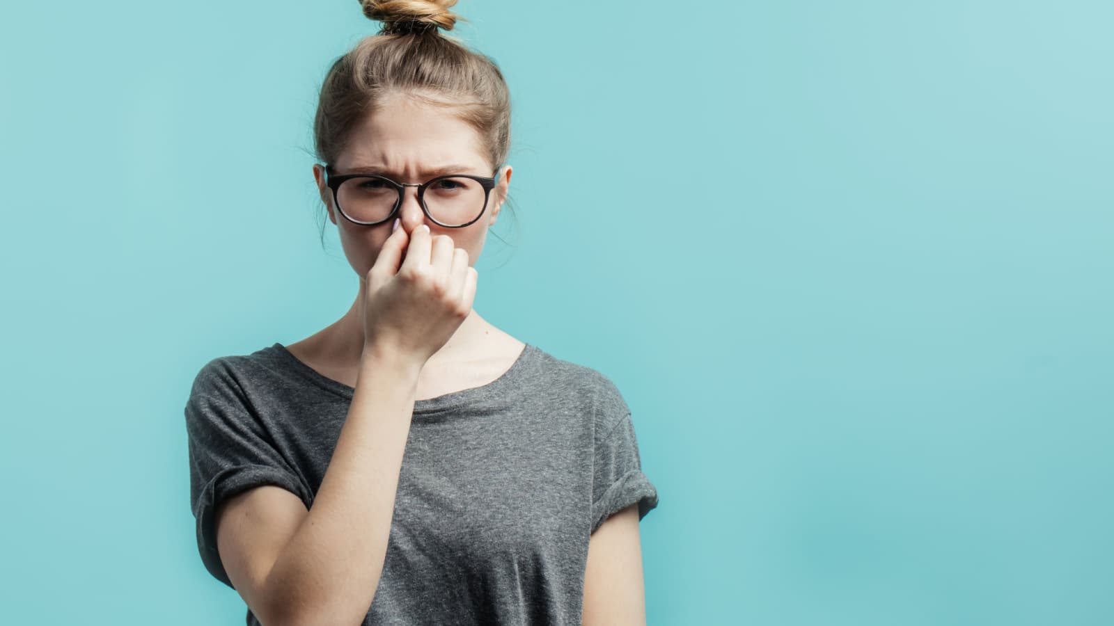 Smells bad, awful odor. Young student girl with hair bun in casual t-shirt squeezing nose with fingers, looking in camera with disgust expression, eager to slip away from bad smell indoors