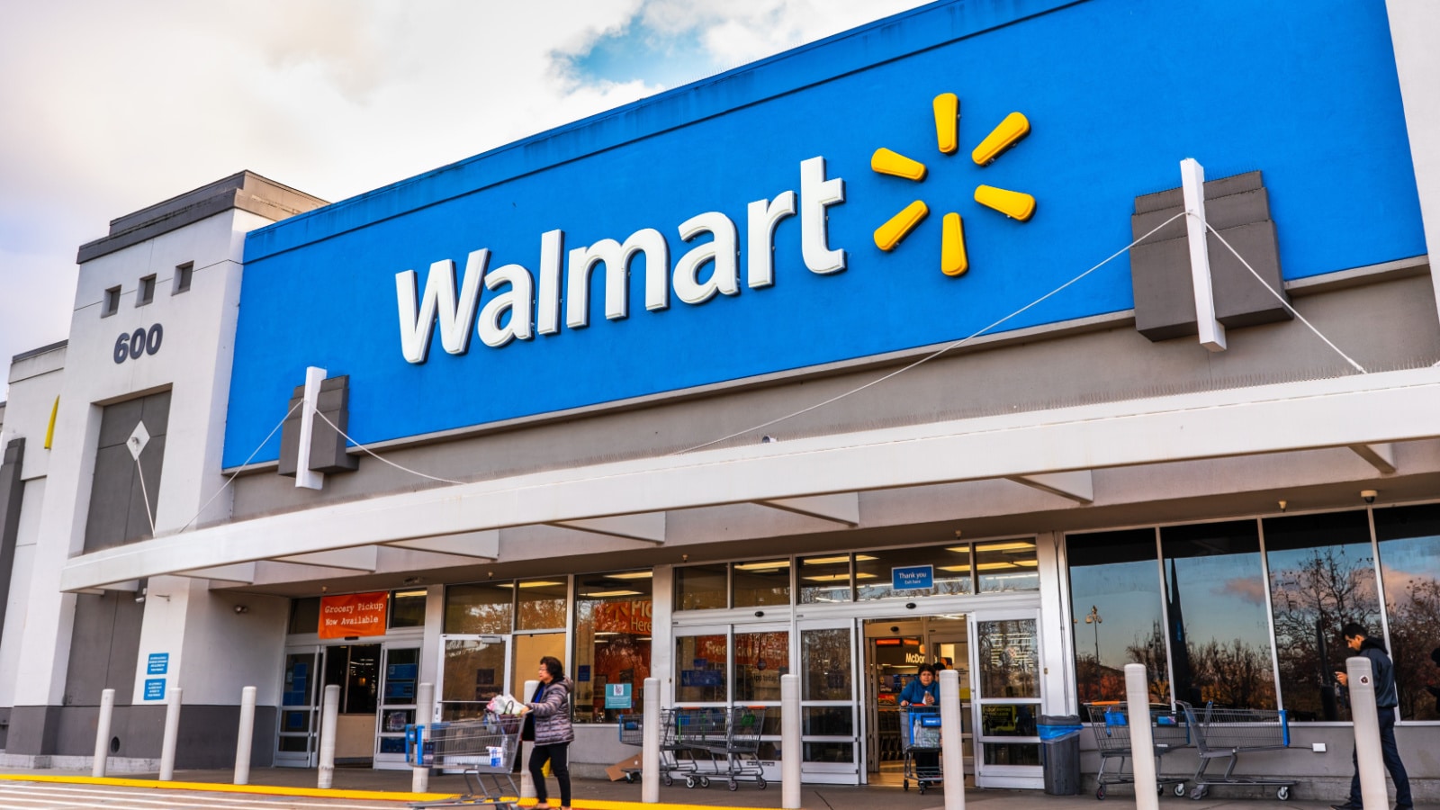 The front entrance of a Walmart store on a sunny day. A woman pushes a cart away from the front entrance doors.