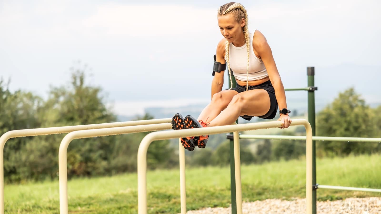 Sporty young female working out calisthenics in an outdoor gym using parallel bars. Beautiful blonde girl with braids works out outdoors doing push-ups on an elevated bar.