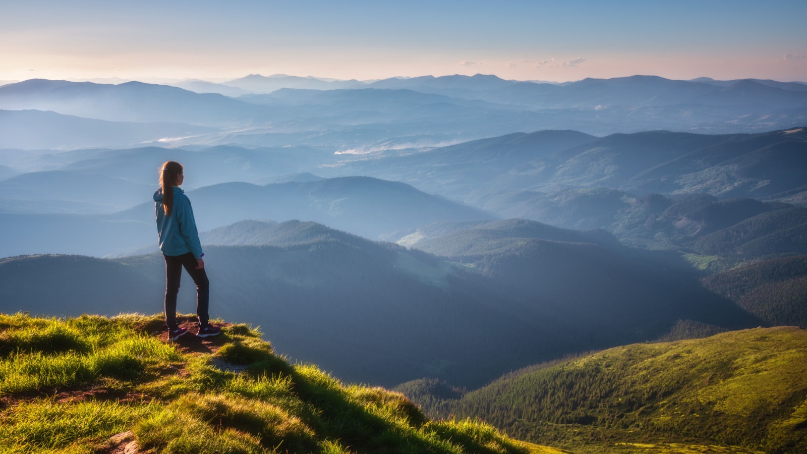 Girl on mountain peak with green grass looking at beautiful mountain valley in fog at sunset in summer. Landscape with sporty young woman, foggy hills, forest, sky. Travel and tourism. Hiking