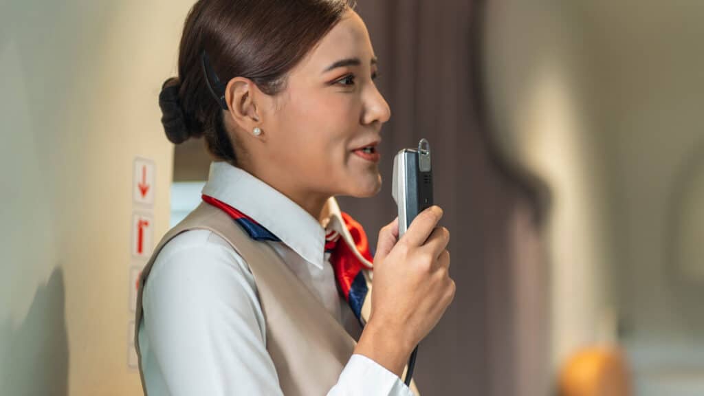 Asian female flight attendant holds a microphone talking to passengers on board an airliner serving passengers during their journey.