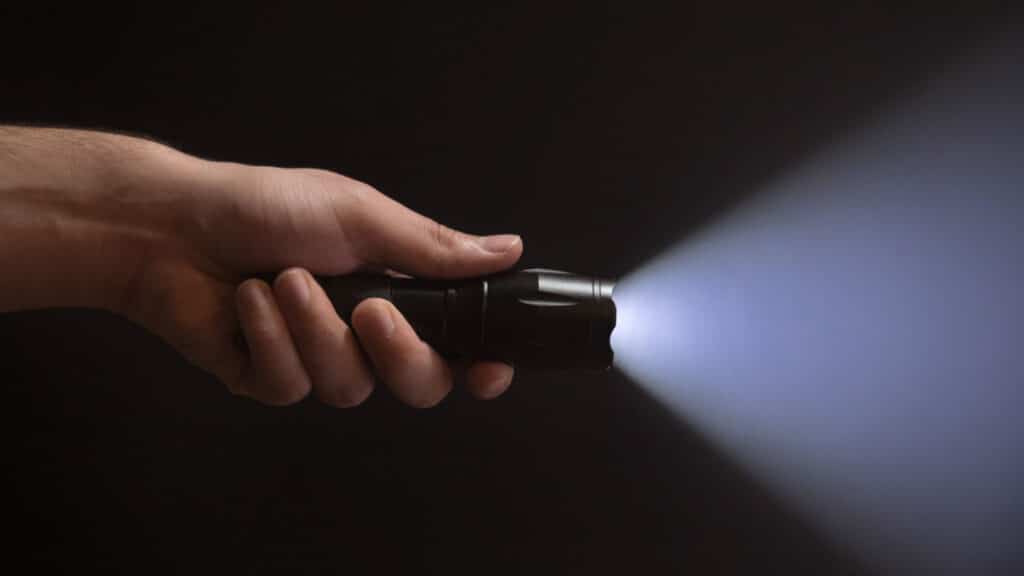 Black flashlight with wide beam in male's hand isolated from left side of the frame on black background