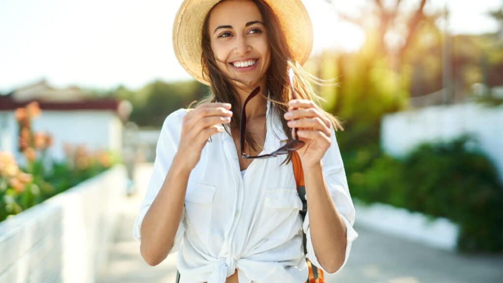 Portrait cheerful tourist woman walking ancient street. Attractive woman in straw hat, white shirt wearing sunglasses, enjoying solo vacation in Europe. Tourism and travel concept.