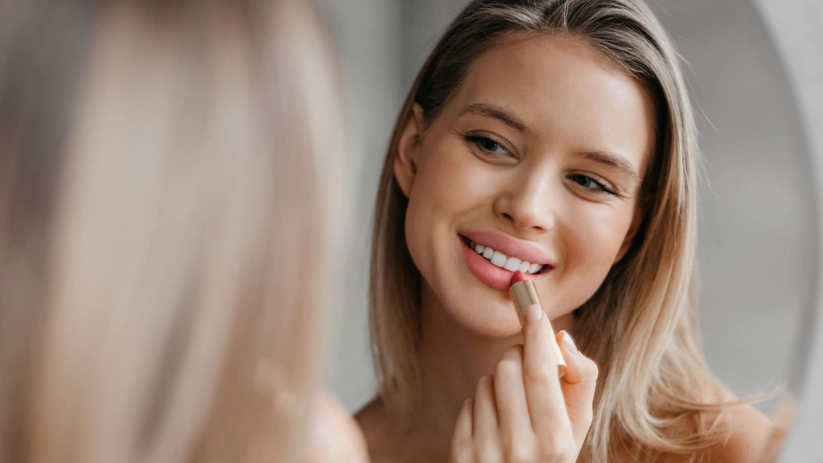 Routine procedures and decorative cosmetics for homemade visage. Happy young woman applying matte lipstick in bathroom interior, looking in mirror, closeup