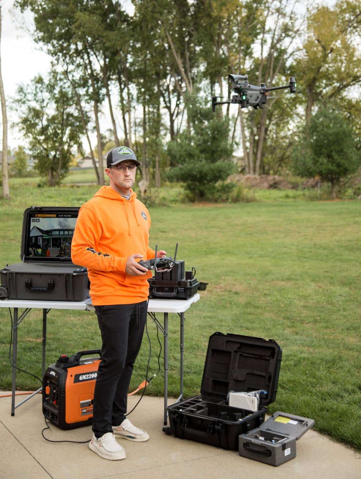 Zac wearing a bright orange sweatshirt with black travel pants and Hey Dude shows, flying a drone with a large monitor behind him