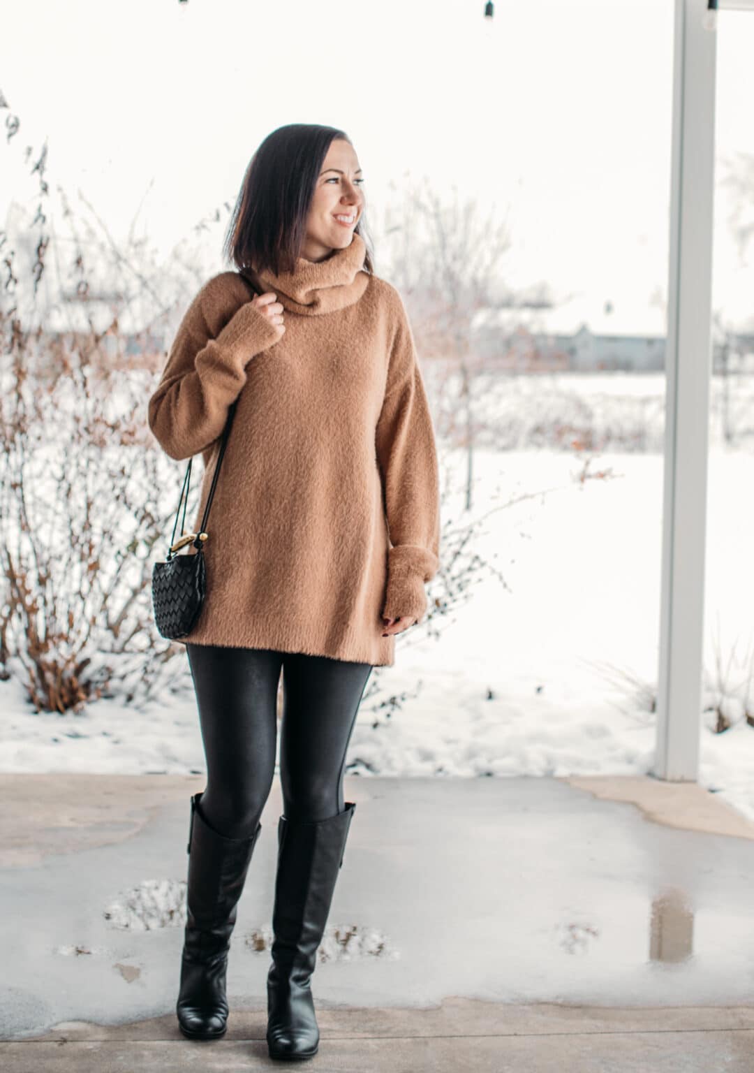 Woman with short dark hair wearing a long tan sweater with leather leggings and black knee high boots in a snowy environment