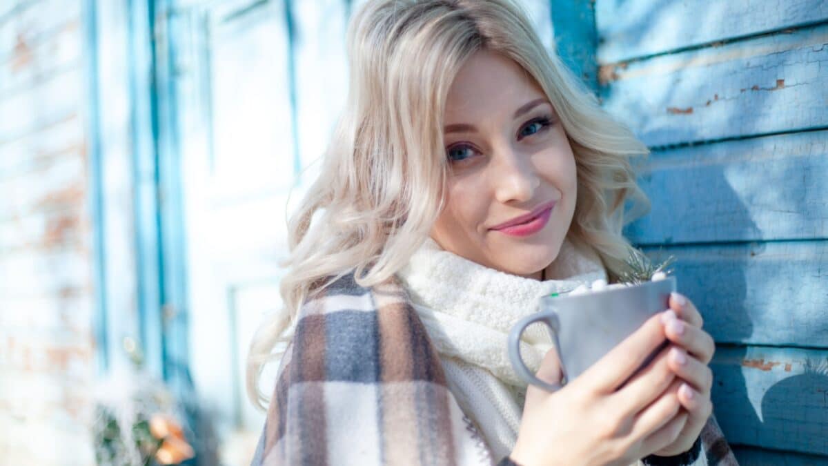 Beautiful blond woman wearing white , sweater, scarf a, hides in a warm brown plaid blanket abd hold the cup against the blue wood wall of log house.