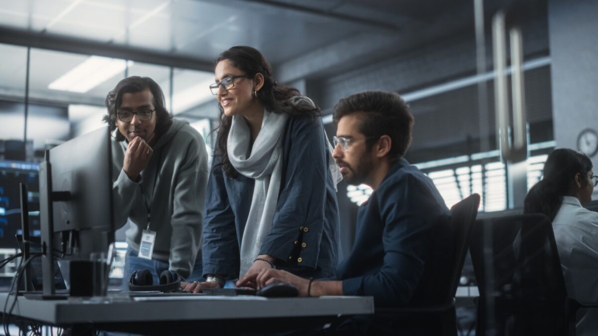 Group of Three Young Indian Software Engineers Use Computer to Discuss a Technological Project in Modern Industrial Office. Group of Male and Female Scientists Work in Research and Development Center