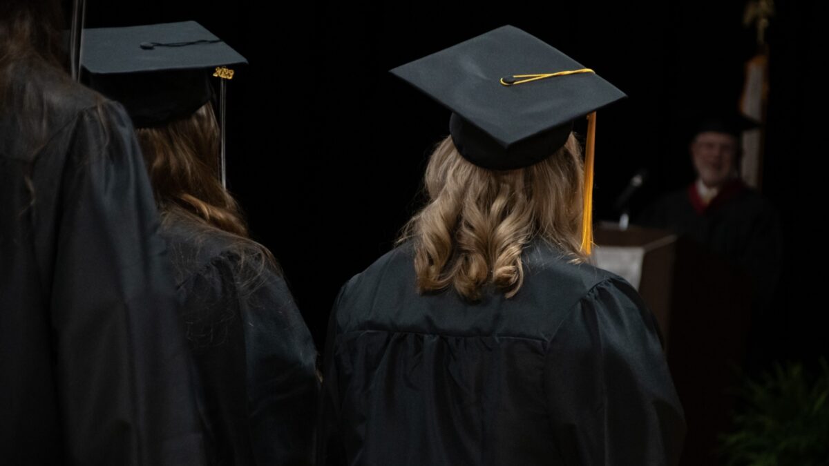 Unidentifiable graduates in black cap, gown, and golden tassel listen to a speech at a community college graduation ceremony.