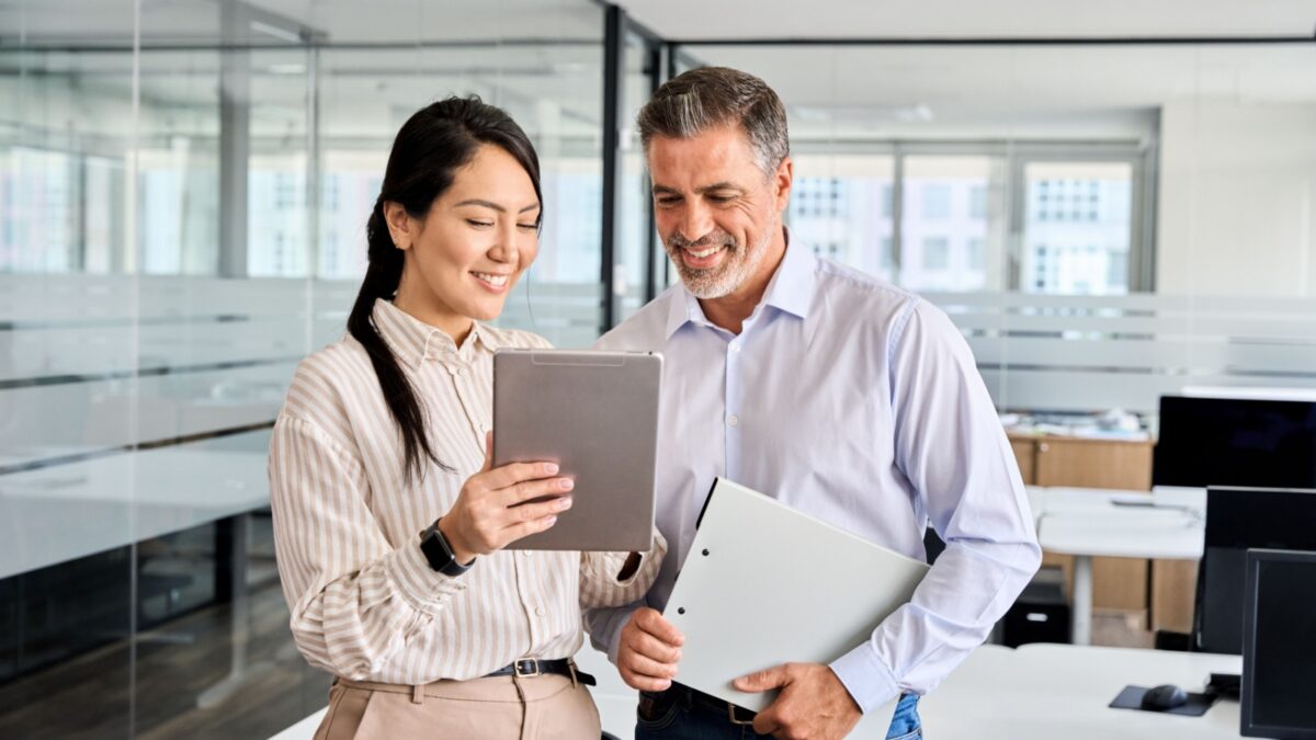 Two happy professional business people team Asian woman and Latin man workers working using digital tablet tech discussing financial market data standing at corporate office meeting.