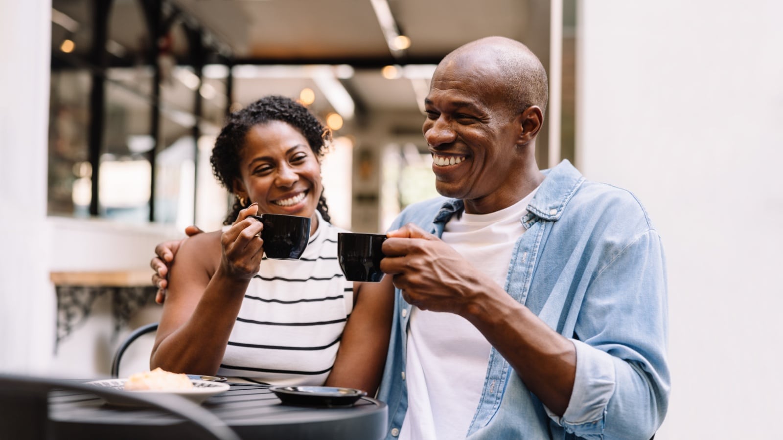 A joyful African American couple share a laugh while sipping coffee in a cozy cafe setting, radiating happiness and warmth.